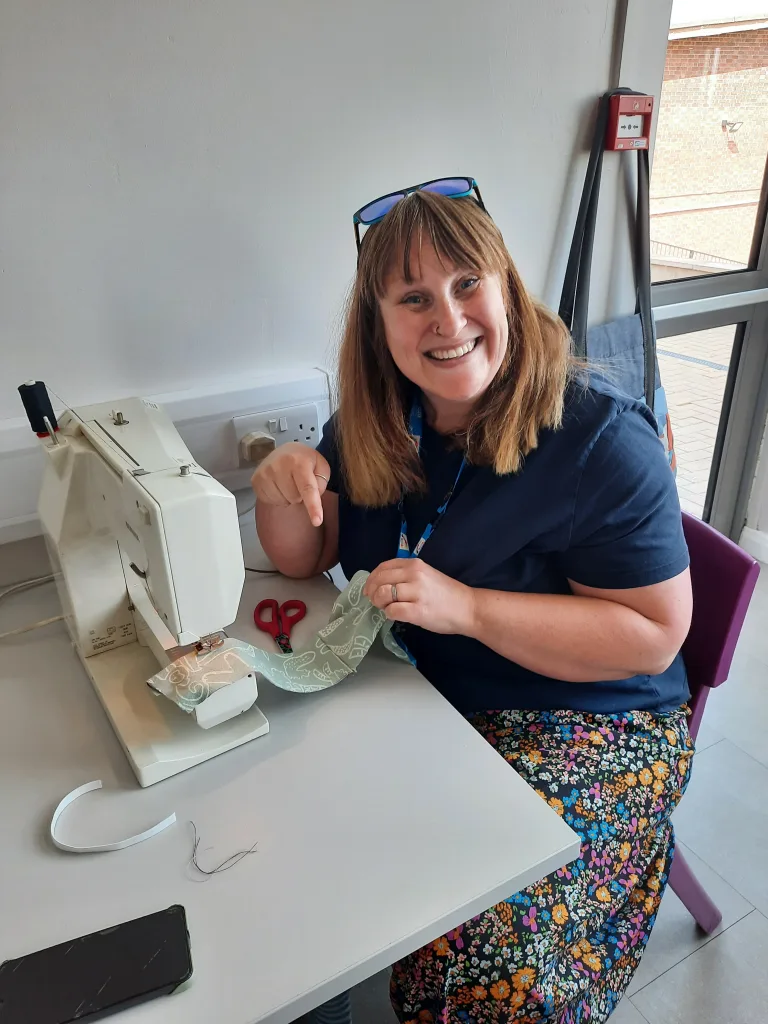 A photo of a woman sat at a sewing machine making a scrunchie, smiling at the camera.