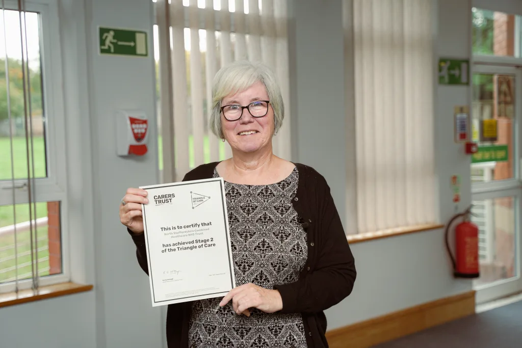Pauline Walsh, Non-Executive Director, standing and smiling, holding a certificate about the Carers Trust accreditation.